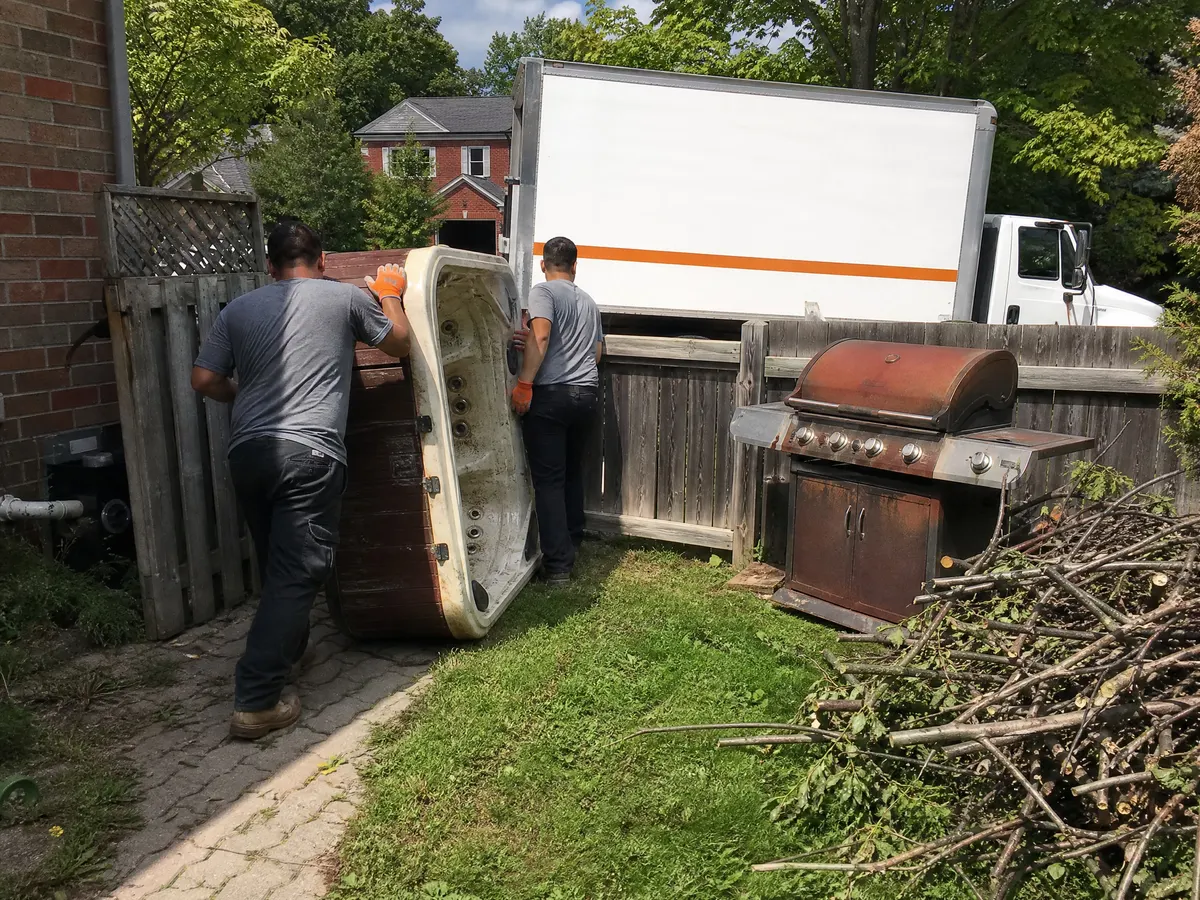Two workers moving an old hot tub through a backyard gate past a rusted barbecue and a pile of branches